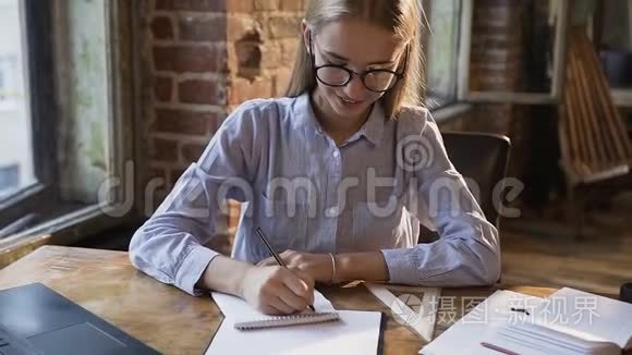 可爱的白种人学生女孩在笔记本上做笔记，坐在红砖旁边的桌子旁学习和工作