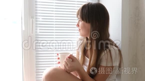 小女孩早上在窗边喝茶。 美丽的女孩看着窗外，手里拿着一杯茶。