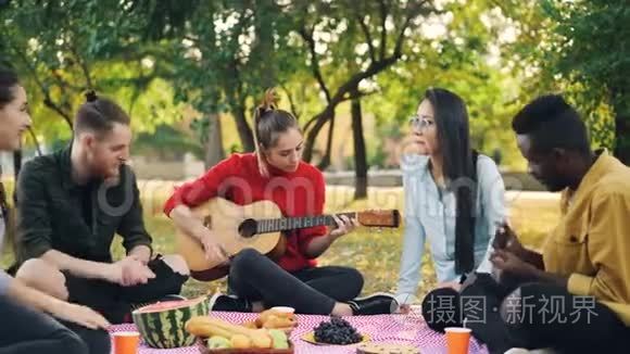 漂亮的女吉他手正在野餐中弹吉他，她的朋友们坐在野餐中唱歌，拍手