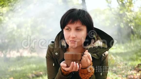 一位女游客在炉火旁的秋天花园里从杯子里喝热茶或咖啡。 漂亮的女孩在发抖