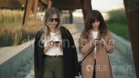 两位美丽的时尚博主在公园散步，在智能手机上上网