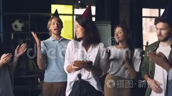 年轻的女士拿着生日蛋糕，吹着蜡烛站在办公室里，同事们鼓掌