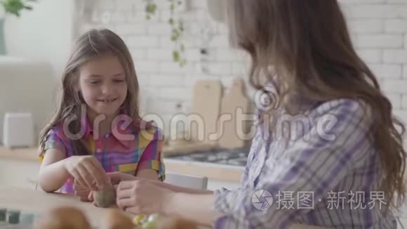 肖像母亲和可爱的女儿坐在复活节的桌子上。 小女孩画了一个带有细流苏的复活节彩蛋