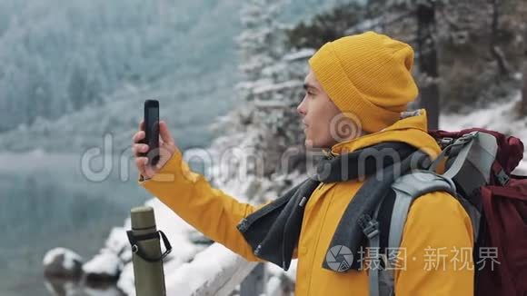 穿着黄色夹克的年轻帅哥站在美丽的山上。 徒步旅行者拍摄冬季自然的视频。 这就是