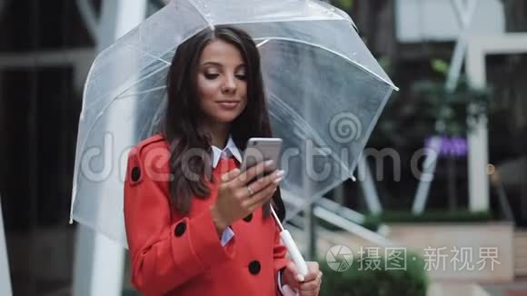 快乐的年轻商务女士站在城市街道上，穿着红色外套使用智能手机。 她拿着雨伞