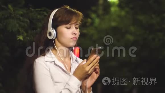 戴着耳机的漂亮女孩在智能手机上听音乐和跳舞。 1.女孩晚上在城市公园散步