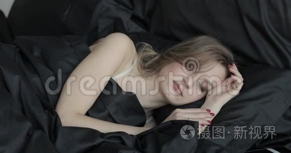 睡在黑色枕头和黑色毯子下的迷人的金发年轻女人的肖像。 她在说话