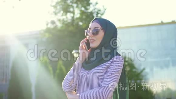 年轻时尚迷人的妇女戴着头巾和眼镜在电话里交谈，阳光明媚。 慢节奏