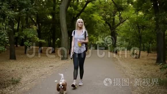 一个长着金发的年轻女人，穿着黑色紧身裤，背着她的宠物带着皮带的骑士走在格雷姆公园旁