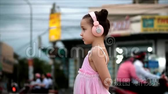 一个可爱的孩子站在路边的肖像，一个可爱的小女孩穿着粉红色的连衣裙和粉红色的耳机看着