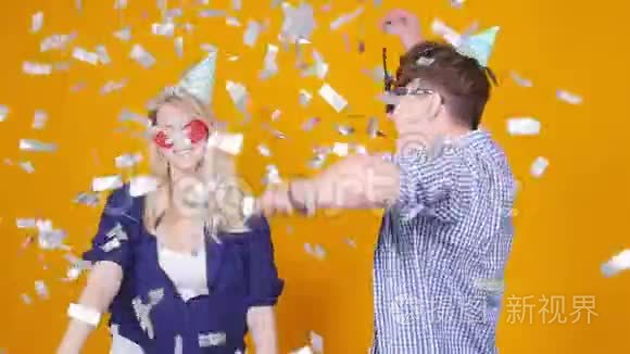 节日和生日的概念。 年轻的幸福夫妇戴着橙色背景的帽子和五彩纸屑跳舞