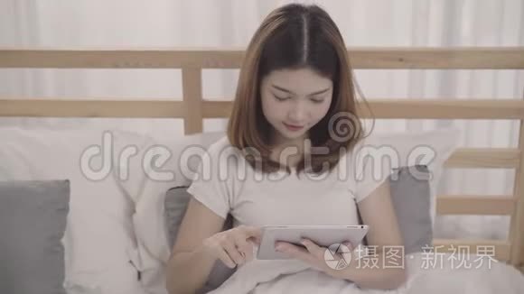 年轻的亚洲女人在早上醒来后躺在床上使用平板电脑。