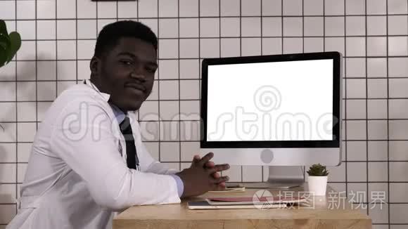 坐在电脑屏幕旁边的黑人医生和摄像机说话。 白色显示器。