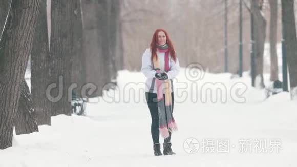 冬季公园。 一个开朗的女人，一头鲜红的头发站在人行道上，做一个雪球，朝前扔去
