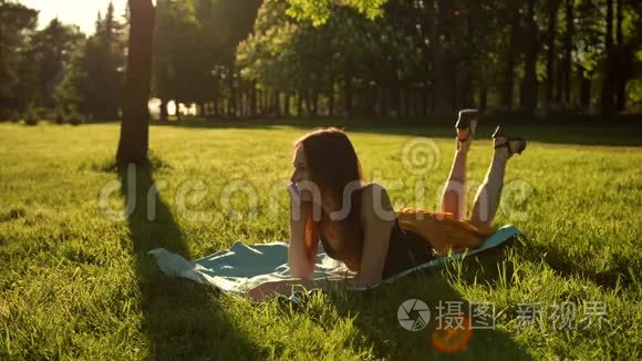 漂亮的女孩躺在草地上，用智能手机说话。 女性从腹部到背部翻转。