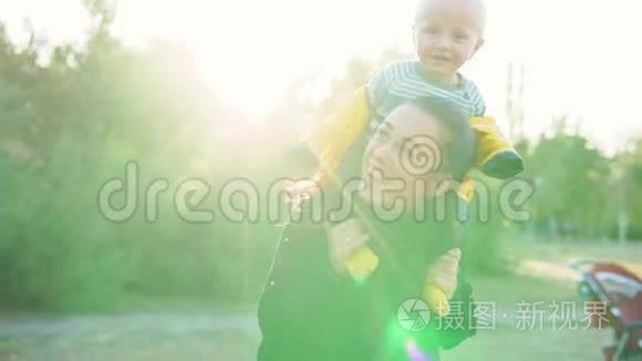 日落时分，妈妈和她的孩子在公园里玩。 男孩穿着一件黄色夹克坐在他的肩膀上，开心地笑着