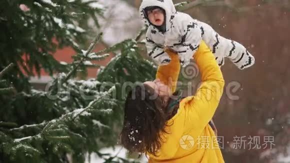 年轻的母亲亲吻她的孩子。一个女人抱着她的小儿子在雪下的冬季公园里散步