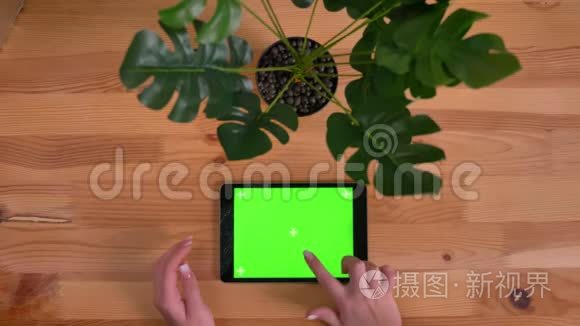 在木桌和植物背景上水平平板的破绿色屏幕上打字的女性手的顶部镜头。