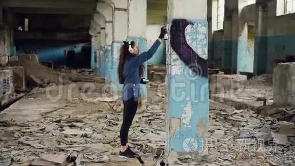城市艺术家正在废弃的仓库里用气溶胶油漆用抽象的图像装饰柱子。 女孩穿着休闲装