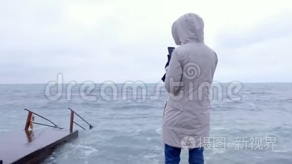 穿白色夹克的女人拿着手机风浪站在堤岸上。 后景。