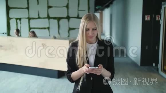 年轻的正式女性在办公室里使用智能手机。 时尚的金发女郎穿着夹克站在当代的办公室大厅里