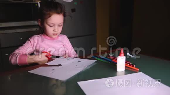 美丽的学龄前欧洲小女孩，穿着粉红色毛衣，独自坐在家里的桌子旁，从纸上剪下形状。