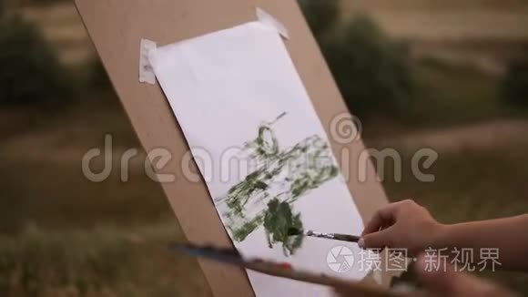 一个可爱的女人用画架和调色板在草地上画户外的侧面视图。 模糊的湖景