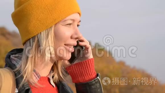 秋天，一个戴着黄色帽子的漂亮白种人女孩正在户外用手机聊天。 微笑的女孩
