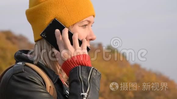 秋天，一个戴着黄色帽子的漂亮白种人女孩正在户外用手机聊天。 微笑的女孩