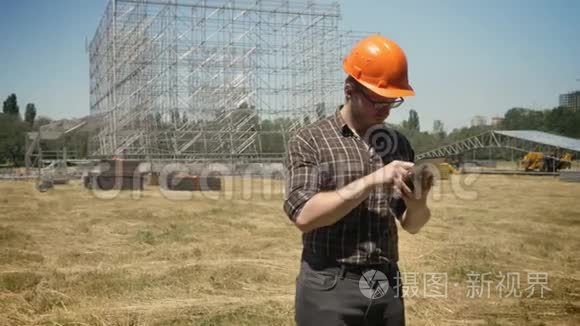 头盔和眼镜的建筑师，使用平板电脑，站在草田上，背景上的金属建筑，集中