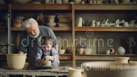 关心的祖父经验丰富的陶工正在教小男孩如何与粘土在陶工`的车轮。 孙子在做
