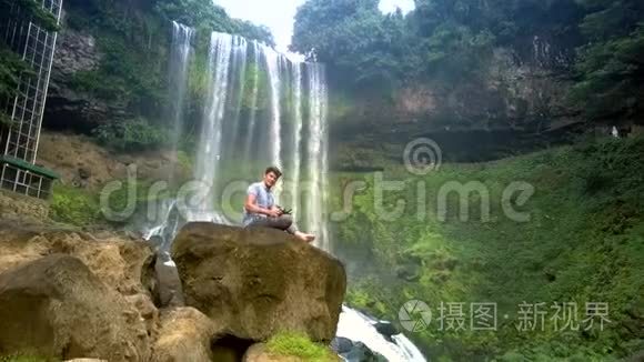 一个人坐在岩石上，用飞摄像头遥控，在瀑布旁微笑