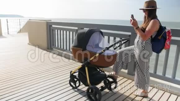 一位年轻的母亲带着婴儿车沿着木板路走，用手机和自拍。 全垒打。
