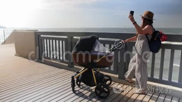一位年轻的母亲带着婴儿车沿着木板路走，用手机和自拍。 全垒打。