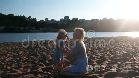 美丽快乐的金发妈妈和女儿拥抱和说话，坐在海滩上日落。