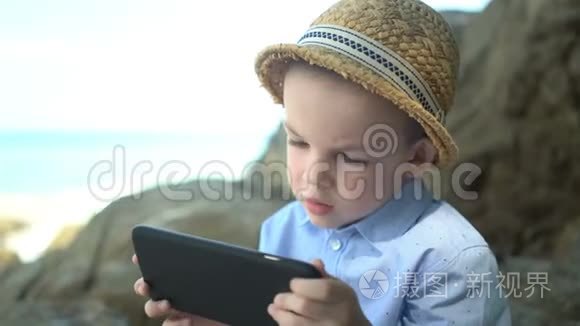 可爱的男孩坐在石头上，使用智能手机，在海边看手机应用程序
