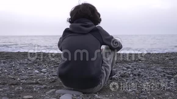 一个孤独的男孩坐在岸上，向大海扔石头。 4k，慢动作