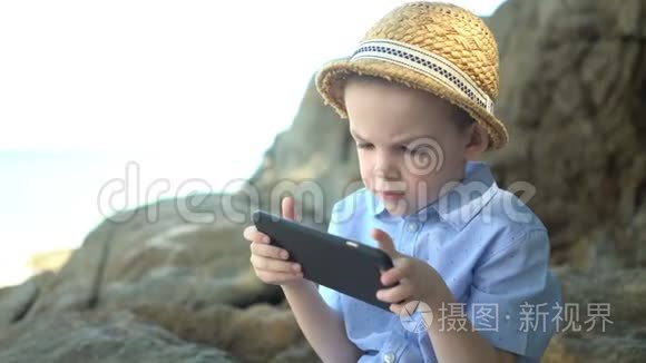 可爱的男孩坐在石头上，使用智能手机，在海边看手机应用程序