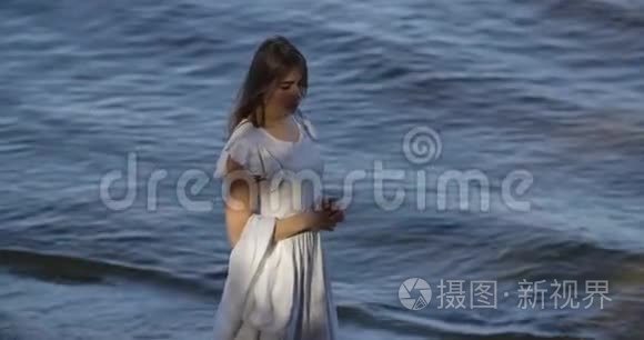 美丽的黑发白种人女人穿着浅色的白色连衣裙站在海浪的背景上思考。 吸引力