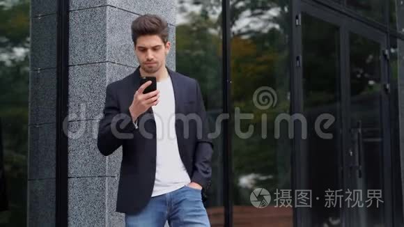 美国帅哥在办公大楼背景下智能手机上使用商务应用程序。 穿西装的年轻商人