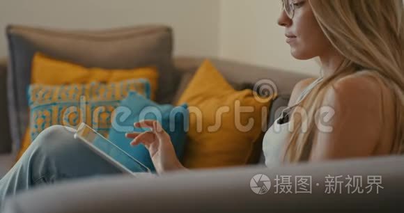 高质量的4k12位镜头，美丽的女孩使用平板电脑坐在家里的沙发上。 女人上网，社交