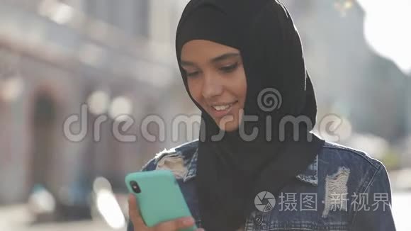 年轻微笑的穆斯林妇女戴着头巾站在市中心，使用智能手机。 通信
