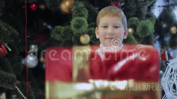 快乐的微笑男孩展示了红色新年礼物。 小孩站在圣诞树下