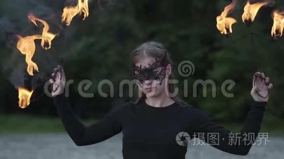 戴着面具的年轻美女在树前用火焰表演节目的肖像。 有技巧的消防艺术家