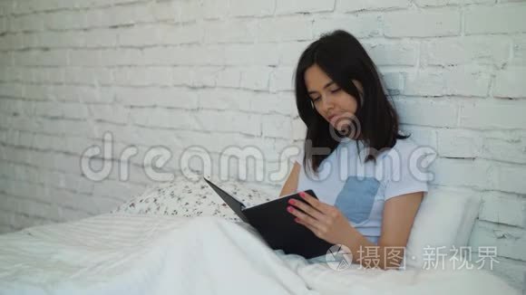 美丽的黑发女孩坐在床上，拿着一本书看书。 一个年轻的女人打开一本有趣的书。