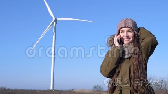 干净的力量，手里拿着手机的女孩微笑着，在现代风车和天空的背景下用电话交谈