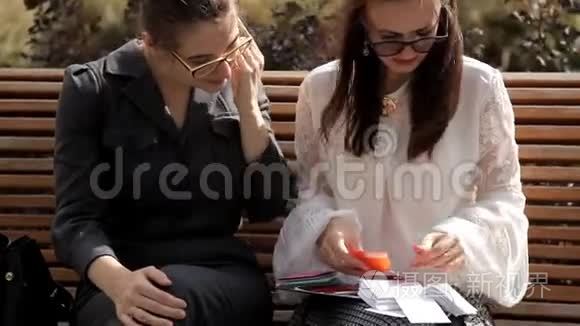 夏天，两个年轻的商务女孩坐在商务中心附近的公园里，考虑彩色卡片
