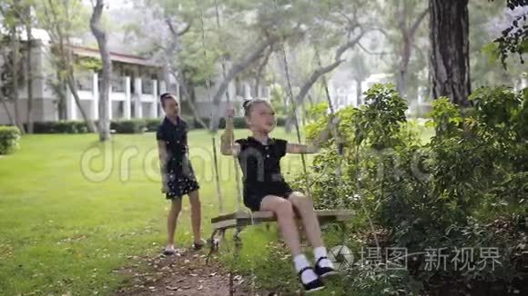 姐姐在阳光下的树下摇着女儿。 女孩笑，高兴。 公园里的家庭乐趣