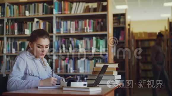 年轻美丽的微笑学生女孩坐在大图书馆的桌子上，有一堆书。 她在写