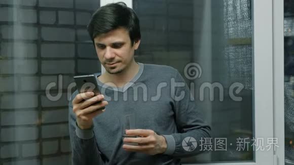 男人正在用手机浏览网页，站在阳台上的窗户附近，喝着水。